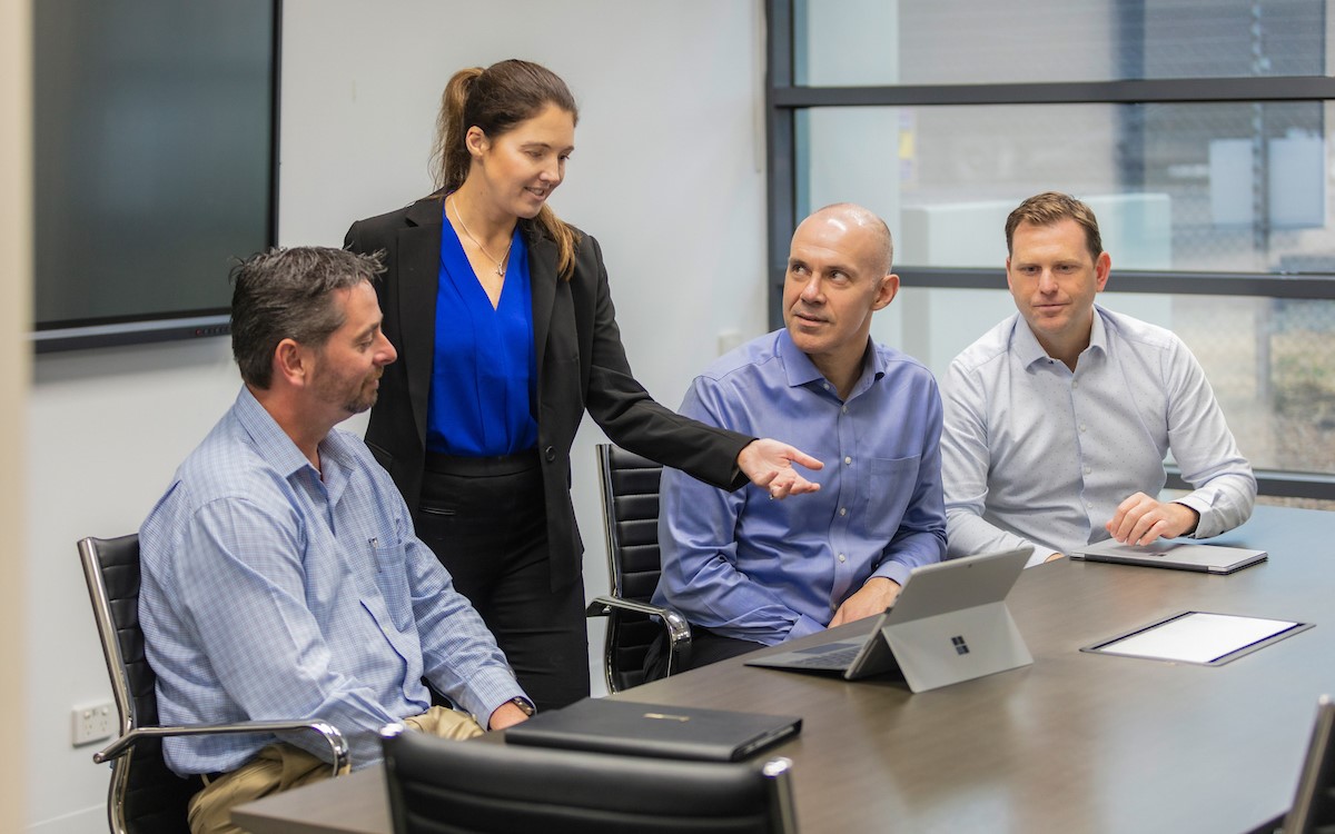 A woman standing and pointing at a laptop screen with three men sitting 