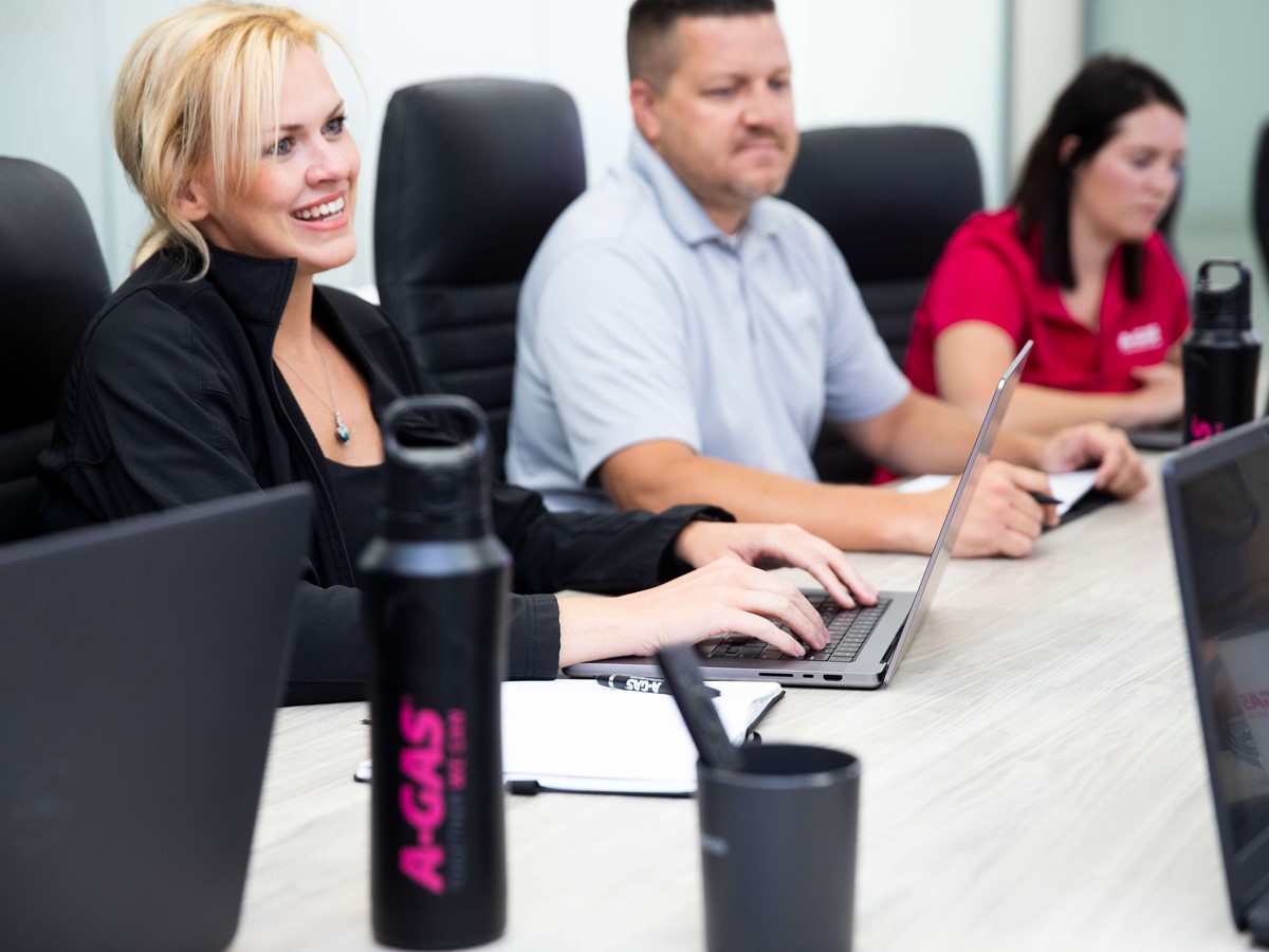 Three people sat at a boardroom desk with laptops, they are all smiling