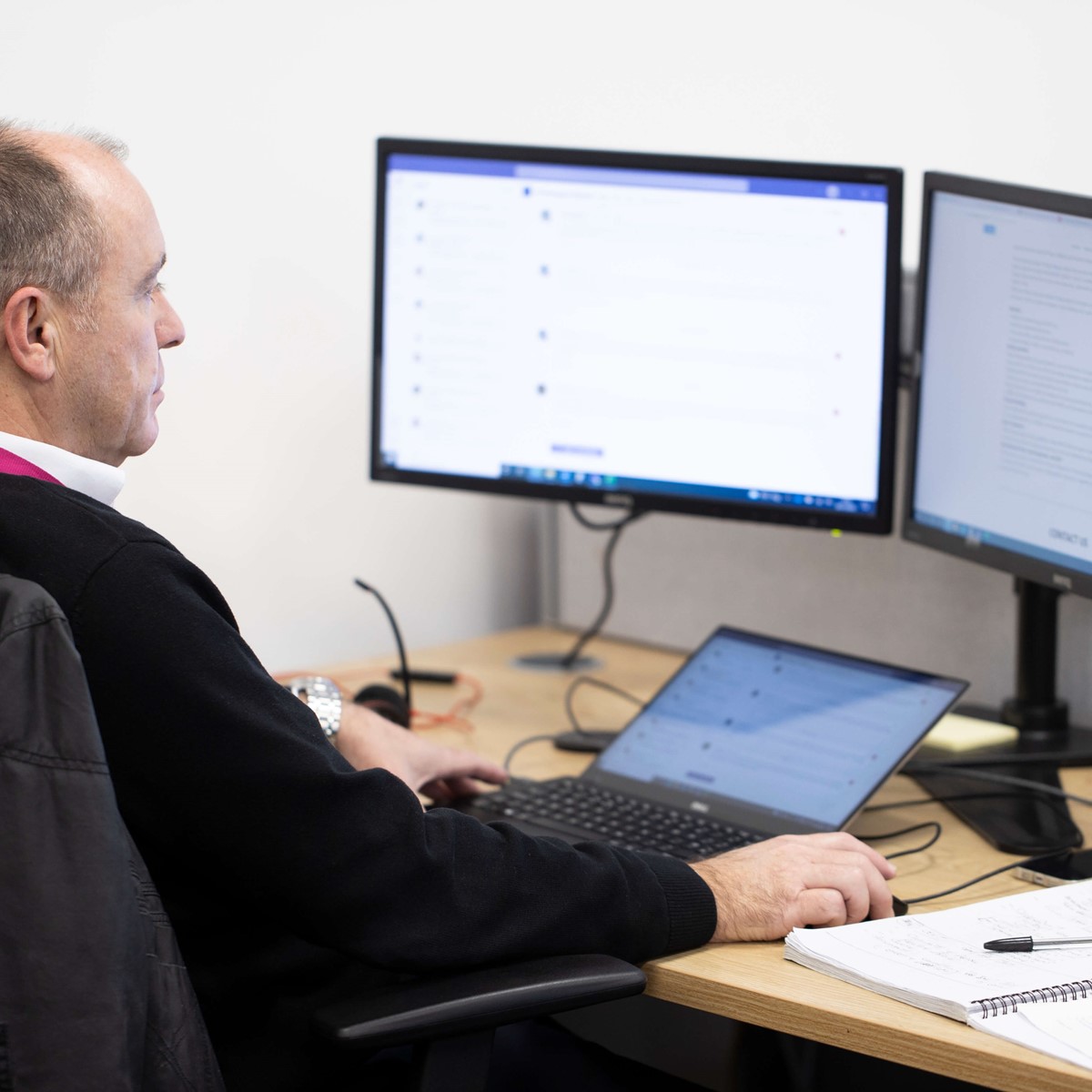 A man sat at his computer screens. The image is taken from behind so we can see him looking at his screens