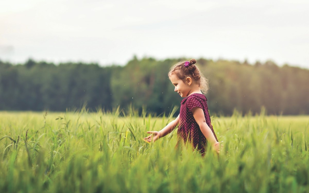 a young girl walking right to left through a green field with forest in background