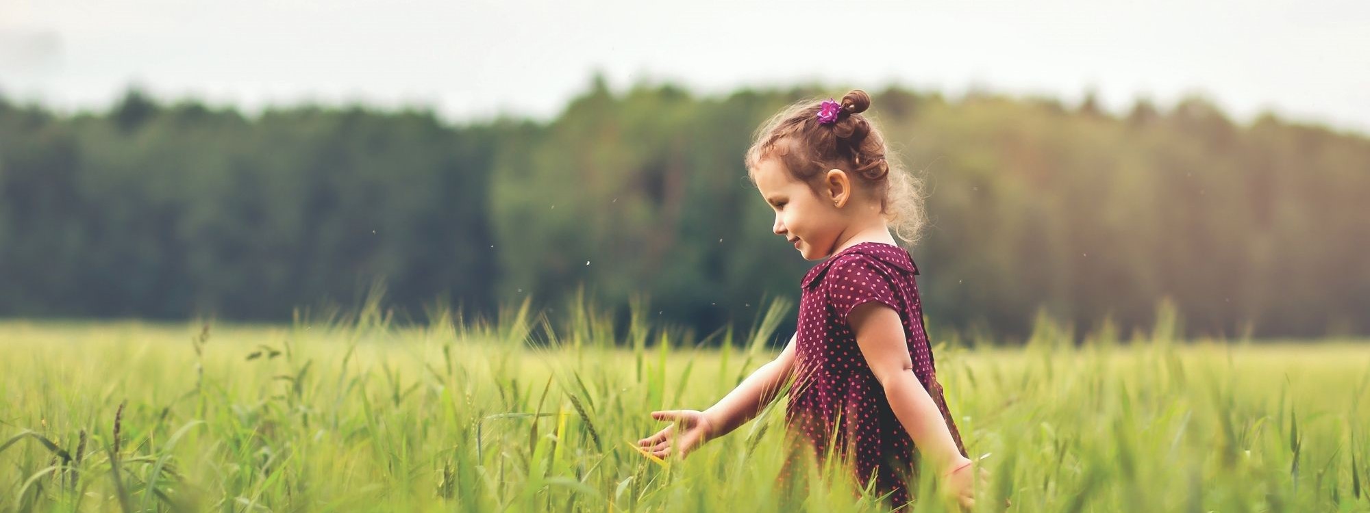 a young girl walking right to left through a green field with forest in background