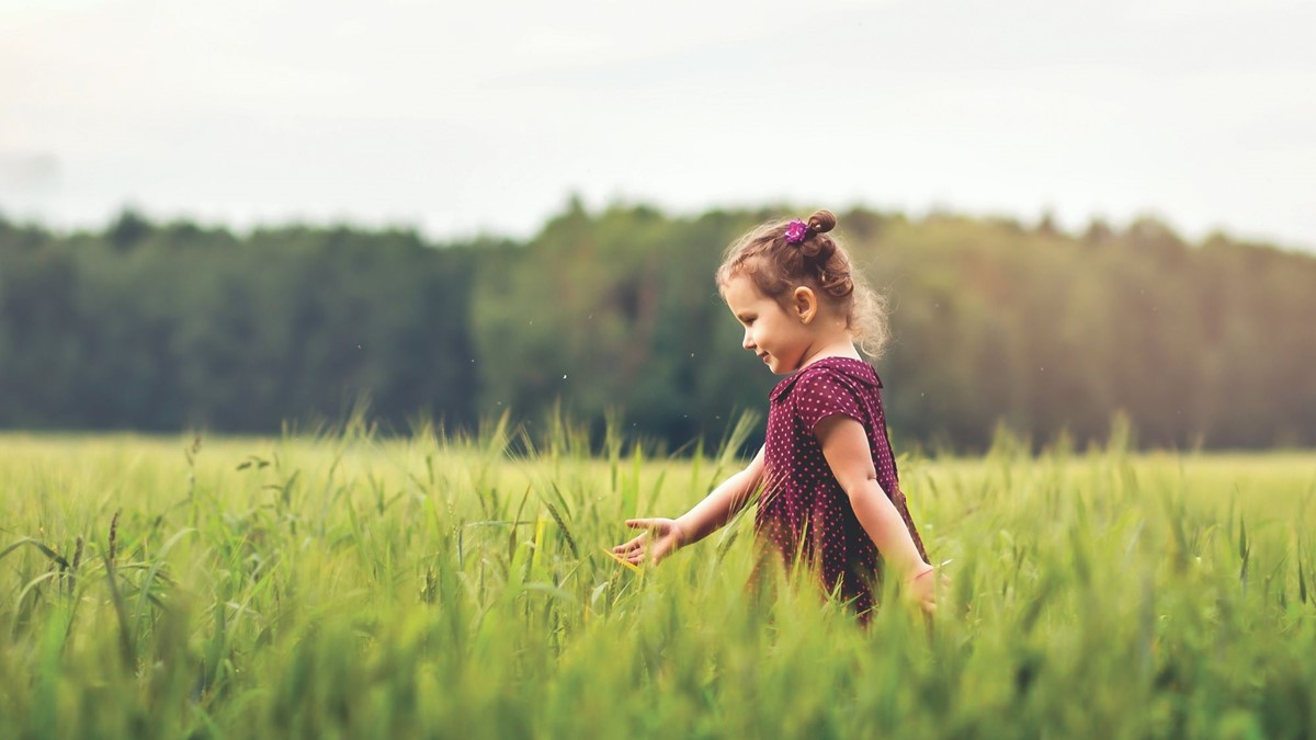 a young girl walking right to left through a green field with forest in background