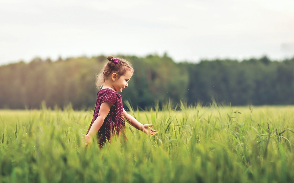 young girl walking left to right through a green field with forest in background