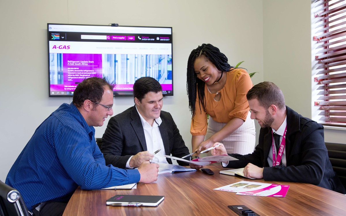 Four people gathered around a table in a meeting, three men are sitting and one woman is standing, they are all looking at a document