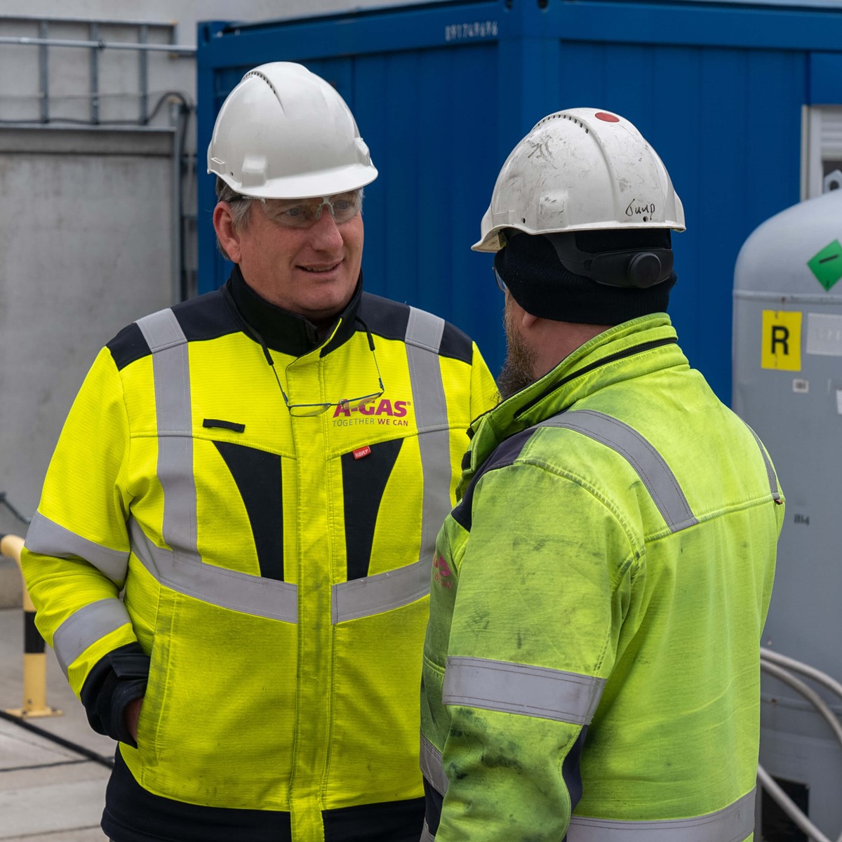 Two men in helmets and yellow hi-vis jackets talk outside 