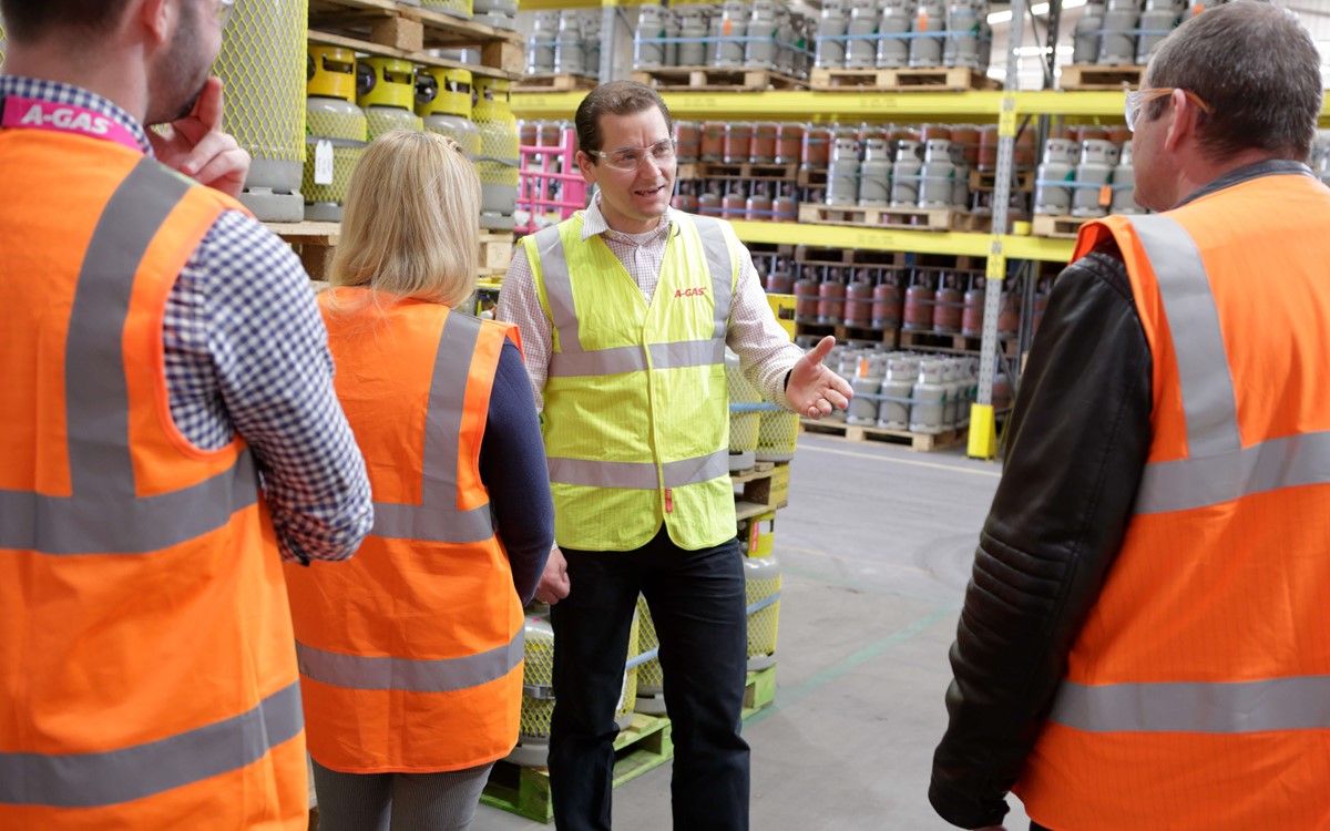 A man in a hi vis giving a site tour to visitors, we can only see the back of visitors heads, the man is surrounded by palettes of gas cylinders