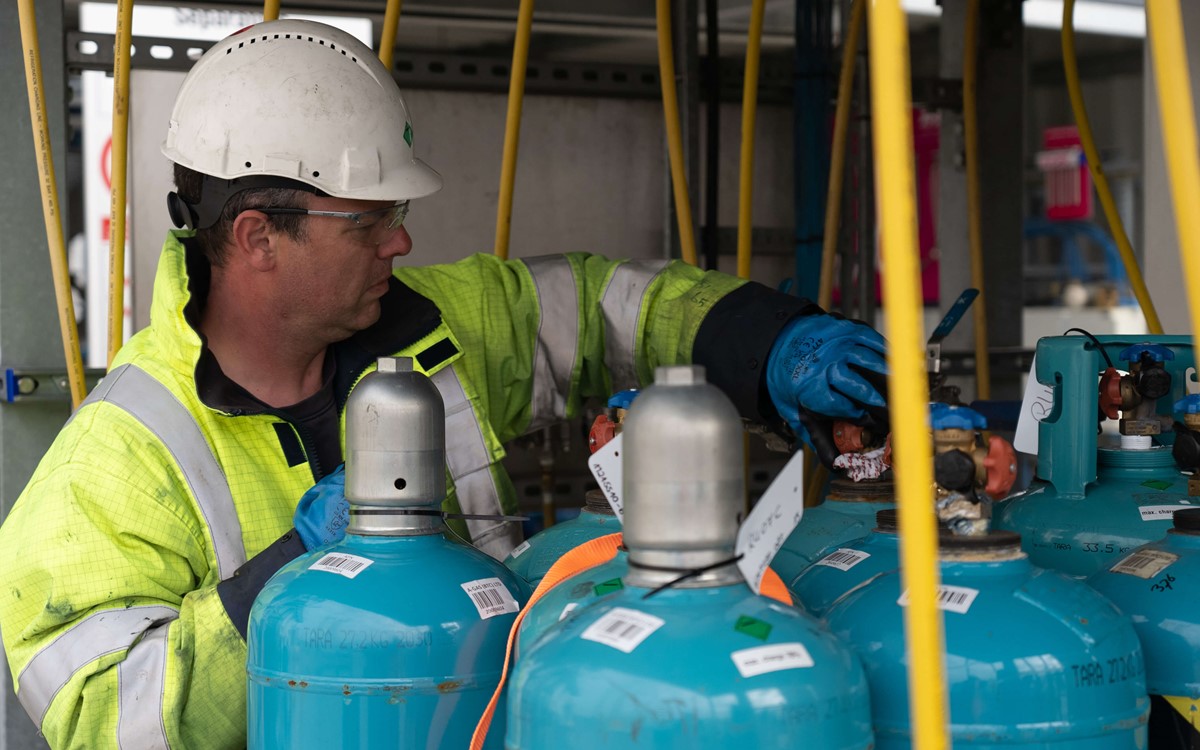 A man in a helmet and yellow hi-vis jacket works on blue cylinders 