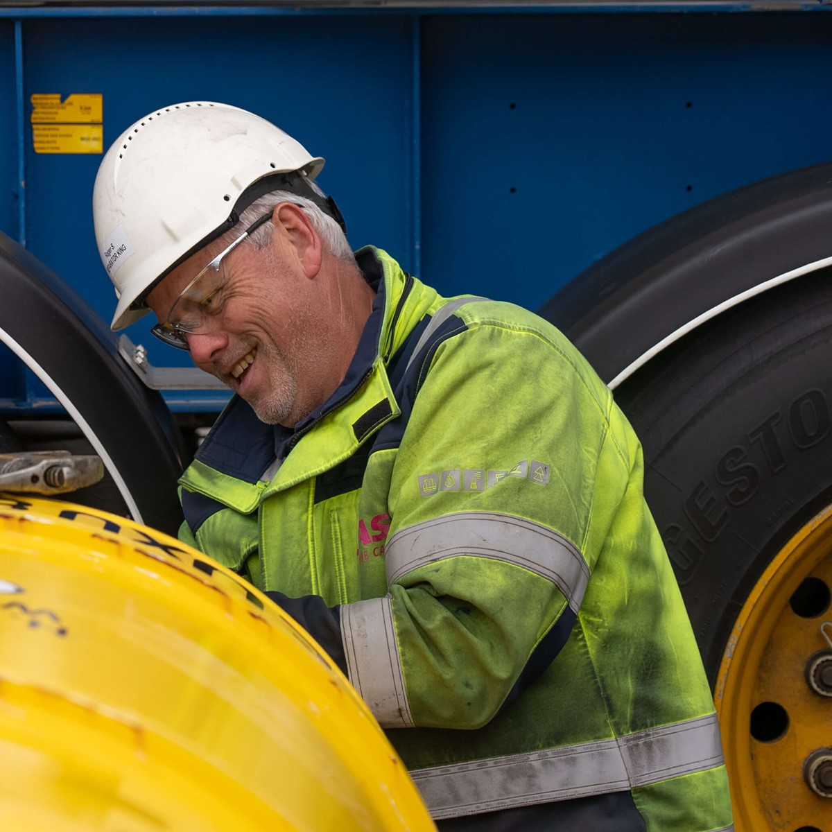 Man in a helmet and yellow hi-vis jacket laughing while working on a yellow tank