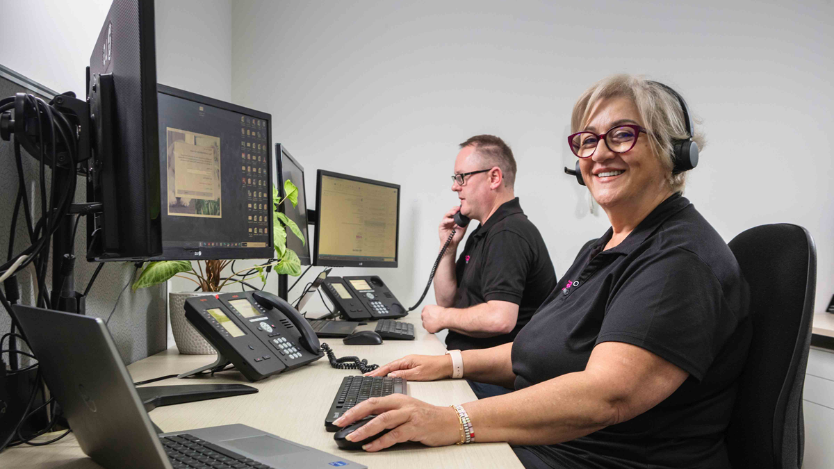 A woman in a headset working at a computer while smiling at the camera 